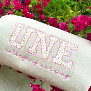 Może przedstawiać: Biała bluza z haftem różowych kwiatów z napisem "ONE college of nursing".