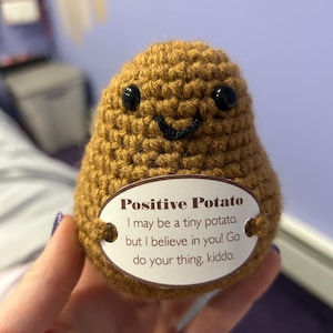 Pode incluir: Uma batata castanha de crochê com olhos pretos e um sorriso costurado. A batata tem uma etiqueta que diz "Positive Potato" com uma mensagem inspiradora. Um item artesanal único e alegre.