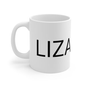 May include: A white ceramic coffee mug with a black sans-serif font spelling out the name 'LIZA' is shown in a studio shot. The mug has a C-shaped handle. The mug is sitting on a white surface.