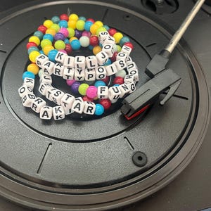 May include: A black record player with a beaded bracelet that spells out "KILL PARTY POISONS FUN" and "BRAK STAR". The bracelet is made of white letter beads and colorful round beads.
