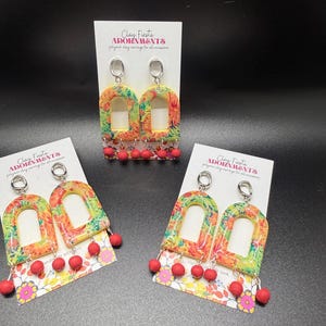 May include: Colorful polymer clay earrings with a floral pattern and red bead accents. The earrings are in an arch shape and hang from silver-colored hooks. The text "Clay Fiesta ADORNMENTS" is printed on the display card.