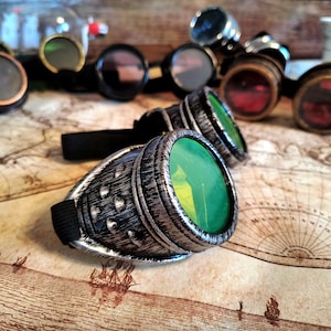 May include: A pair of steampunk goggles with green lenses and silver-colored metal frames. The goggles are lying on a map with a compass design.