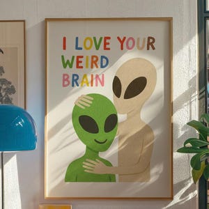May include: A framed art print featuring two alien figures. One green alien is embraced by a beige alien. The text "I LOVE YOUR WEIRD BRAIN" is written in colorful letters above the aliens. The artwork is displayed on a wall.