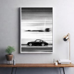 May include: Black and white framed print featuring a classic black sports car and a jet plane in motion. The artwork has a blurred background, creating a sense of speed and movement. The print is mounted in a white frame.