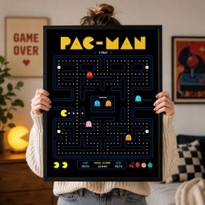 May include: A framed Pac-Man arcade game poster featuring a black background with a yellow title, a blue maze layout, white dots, and colorful ghost characters. The bottom section displays the text 1UP, HIGH SCORE, 2UP, and various fruit icons.