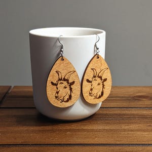 May include: Wooden teardrop earrings featuring a laser-etched goat head design. The earrings are light brown with silver-colored ear wires. The earrings are displayed in front of a white mug.