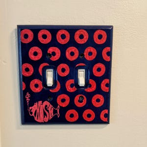 May include: A light switch cover with a blue background and a pattern of red donuts. The cover has two white light switches and a pink fish with the word "FISH" written on it.
