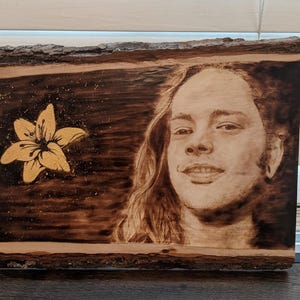 May include: Wood-burned art piece featuring a portrait of a person with long hair and a smiling expression. A golden flower is depicted on the left side. The artwork is on a rectangular wooden board with a natural bark edge.