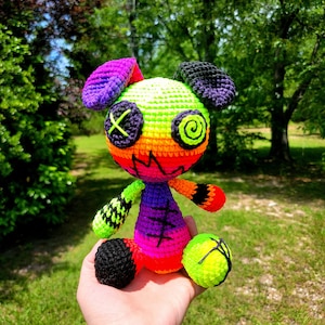 Può includere: Un animal di peluche all'uncinetto colorato a forma di cane con filato arcobaleno, verde e nero. Il cane ha una X cucita per gli occhi e una spirale per il naso.