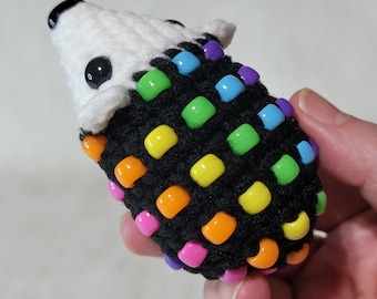 May include: A small crocheted hedgehog figurine featuring a white yarn head with black plastic eyes and a black nose. The body is crafted from black yarn and is decorated with numerous colorful plastic pony beads in shades of orange, yellow, green, blue, and pink.