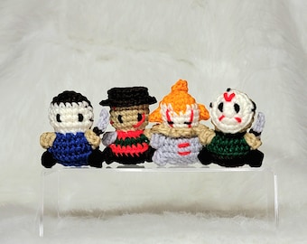 May include: Four crocheted horror movie character figures. The figures are standing on a clear acrylic display stand. The figures are from left to right: a blue and white figure, a red and white figure, a white and orange figure, and a green and white figure.