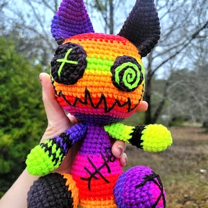 May include: A crocheted plush toy in the shape of a creature with rainbow stripes, black eyes, and a stitched-on smile. The toy has purple ears and black feet.