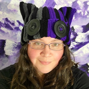 May include: A handmade crochet hat with a cat ear design. The hat features alternating stripes of black, gray, and purple. Large black buttons are sewn onto the front. The hat is worn by a person wearing glasses.