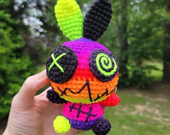 May include: A colorful crocheted bunny with a rainbow pattern, black eyes with an X and a spiral, and a stitched-up mouth. The bunny has a green and yellow ear and a black ear.