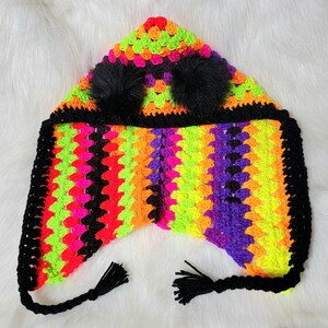 Può includere: Un cappello all'uncinetto colorato con un bordo nero all'uncinetto e due pompon neri. Il cappello è realizzato con filato verde neon, arancione, rosa, viola e giallo.