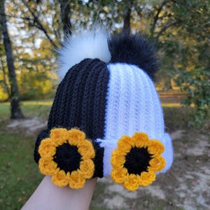 Puede incluir: Un gorro de ganchillo en blanco y negro con un diseño de girasol. El sombrero presenta un diseño de bloques de color blanco y negro, con un pompón blanco y negro en la parte superior. Dos aplicaciones de girasoles amarillos y negros están en el borde.
