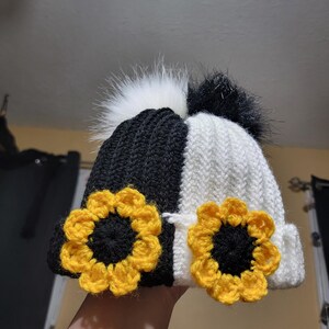 Puede incluir: Un gorro de punto en blanco y negro con un dise&ntilde;o de girasol. El gorro presenta un dise&ntilde;o de color dividido, con una secci&oacute;n en blanco y negro. Dos girasoles de ganchillo amarillos y negros est&aacute;n en la parte delantera. El gorro tiene dos pompones de piel sint&eacute;tica.