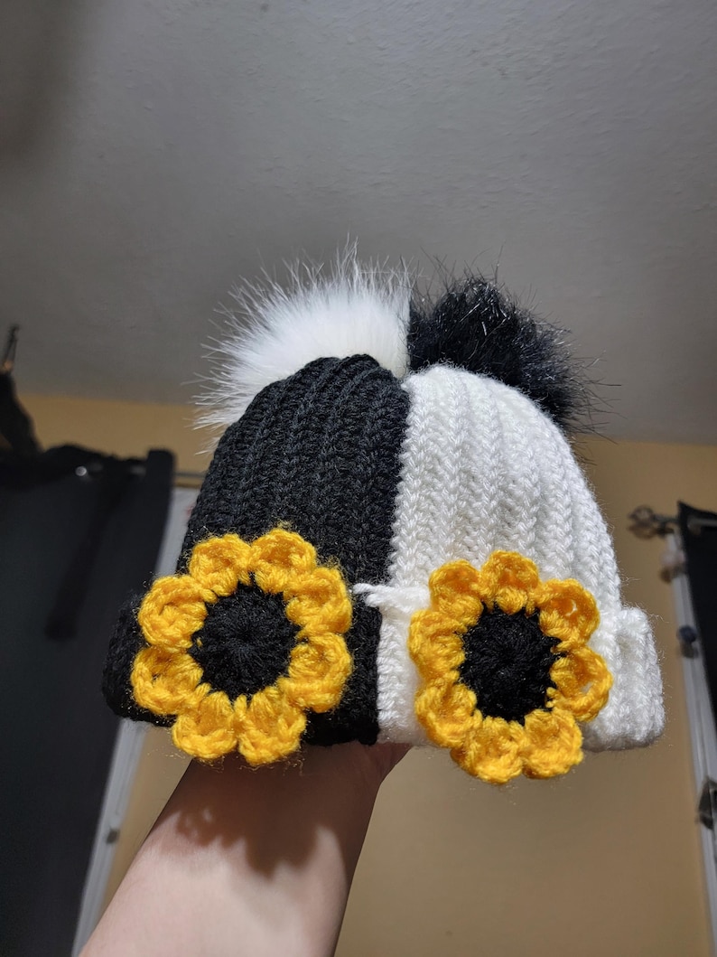 Puede incluir: Un gorro de punto en blanco y negro con un dise&ntilde;o de girasol. El gorro presenta un dise&ntilde;o de color dividido, con una secci&oacute;n en blanco y negro. Dos girasoles de ganchillo amarillos y negros est&aacute;n en la parte delantera. El gorro tiene dos pompones de piel sint&eacute;tica.