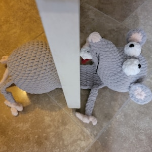 May include: A gray crocheted rat toy with large white eyes and pink ears. The rat is lying on a tiled floor with its head peeking out from behind a white door.