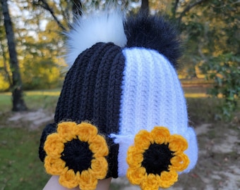 May include: A handmade black and white beanie with a sunflower design. The hat features a black and white color block design, with a white and black pom-pom on top. Two yellow and black crochet sunflowers are attached to the brim.