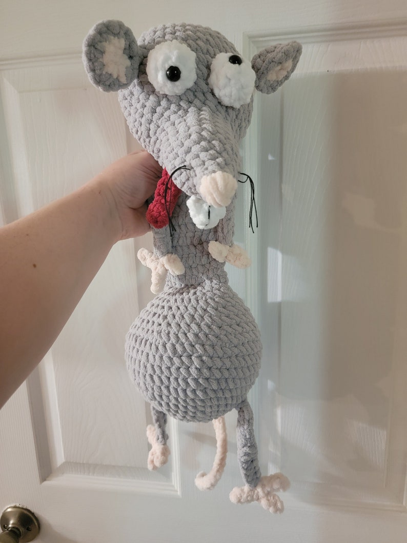 May include: A gray crocheted rat plush toy with large white eyes and a red scarf. The rat has a long tail and is standing on its hind legs.
