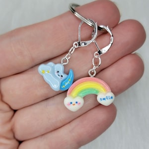 Pode incluir: Um par de brincos extravagantes com uma nuvem azul e um arco-íris. A nuvem tem um rosto sorridente e estrelas amarelas. O arco-íris tem uma base em forma de coração com a palavra "smile" e um rosto sorridente.