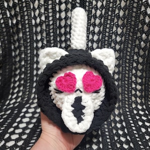 May include: A handmade crochet cat skull with pink heart eyes, black eyelashes, and a black hood. The cat skull is white with a black nose and mouth. A white, cylindrical shape extends from the top of the skull.