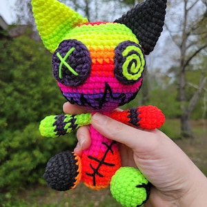 Può includere: Un piccolo giocattolo all'uncinetto a forma di gatto con uno schema di colori arcobaleno. Il giocattolo ha occhi neri con una X e un disegno a spirale, e una bocca cucita. Il giocattolo è fatto con colori al neon luminosi, tra cui rosa, verde, giallo, arancione e rosso.
