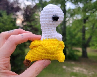 May include: A crocheted banana-shaped plush toy with a white head and a single black eye. The toy is held in a hand against a blurred background of green trees.