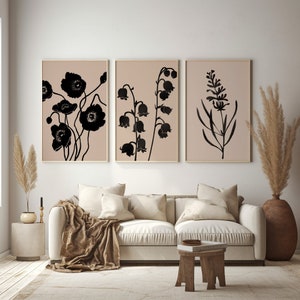 May include: Three black and white floral prints with a light brown background. The prints are framed in a thin, light brown frame. The prints are hanging on a light beige wall above a white couch with brown throw pillows and a brown throw blanket.