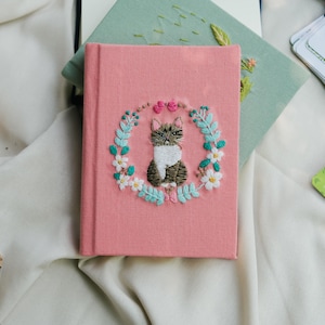 Può includere: Un diario rosa con copertina rigida con un disegno di gatto ricamato. Il gatto è circondato da una ghirlanda di foglie blu, fiori bianchi e accenti rosa. Altri diari e carte sono sullo sfondo.