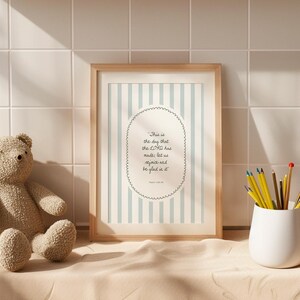 Puede incluir: Una impresión enmarcada con un marco de madera clara presenta un fondo a rayas azul claro y blanco. La impresión incluye el texto "This is the day that the LORD has made, let us rejoice and be glad in it" en un óvalo decorativo. Un oso de peluche y un portalápices blanco también están en la imagen.