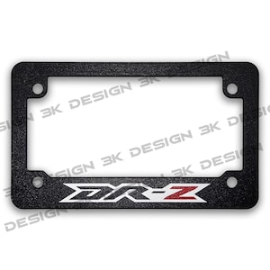 May include: Black license plate frame with the text "DR-Z" in white and red. The frame has a textured surface and mounting holes in each corner. The words "3K DESIGN" are printed around the frame.
