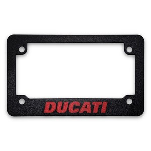 May include: Black license plate frame with the red word "DUCATI" across the bottom. The frame has a textured surface and four mounting holes. The frame is designed to hold a standard license plate.