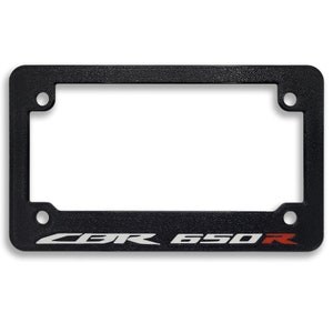 May include: Black license plate frame with the text "CBR 650R" in white and red. The frame has a textured surface and four mounting holes, designed for a motorcycle.