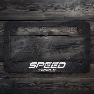Puede incluir: Marco de matr&iacute;cula negro con las palabras "SPEED TRIPLE" en blanco. El marco tiene una superficie texturizada y es rectangular con esquinas redondeadas. El fondo es una veta de madera oscura.