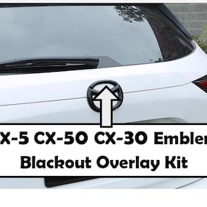 May include: Black emblem overlay kit for CX-5, CX-50, and CX-30 models. The image shows the rear of a white car with a black emblem and the text "CX-5 CX-50 CX-30 Emblem Blackout Overlay Kit."