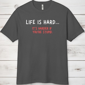 May include: Dark grey t-shirt featuring the text "LIFE IS HARD... IT'S HARDER IF YOU'RE STUPID." in white and red. The shirt has a crew neck.