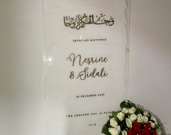 Pode incluir: Uma placa de acrílico branca com letras douradas. A placa diz "Bem-vindos" em árabe, "Soyez les bienvenus" em francês, "Nesrine & Sidali", "23 de dezembro de 2023", e "We created you in pairs 77:8".