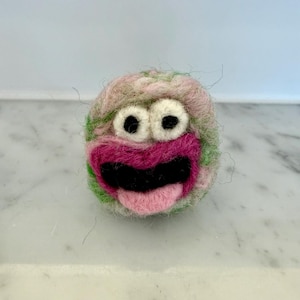 May include: A whimsical, round felted creature with a pink, green, and beige color scheme. It features large white eyes with black pupils, a pink mouth, and a black tongue. The handmade design has a playful, cartoonish aesthetic.