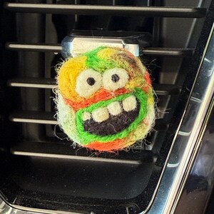 May include: A colorful, handmade air vent clip featuring a whimsical, cartoonish design. The clip has a round shape with a smiling face, showcasing a combination of yellow, green, orange, and black colors. It is designed to attach to a car's air vent.