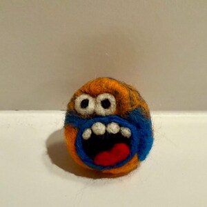 May include: A handmade felted monster ball with orange, blue, and black colors. The monster has white teeth, a red tongue, and wide open mouth. The eyes are white with black pupils. The ball is a whimsical, handcrafted item.