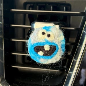 May include: A round, felt air freshener with a cartoon face design. The face features a blue mouth, black teeth, and large, off-white eyes. The body is a mix of blue, gray, and white felt, attached to a car vent.