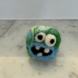 May include: A handmade felt monster ball with a blue, green, and white color scheme. The monster has large white eyes with black pupils, a blue mouth, and a row of black teeth. The ball is round and has a whimsical design.