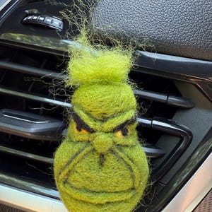May include: A lime green felt air freshener shaped like a character's face with a grumpy expression. The air freshener has black eyebrows and a black mouth. The air freshener is attached to a car vent.