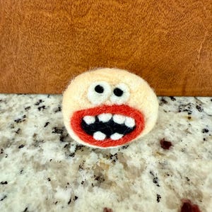 May include: A whimsical, handmade felted character with a light beige body, black eyes, and a wide red mouth revealing white teeth. The felted figure is a unique, handcrafted item.