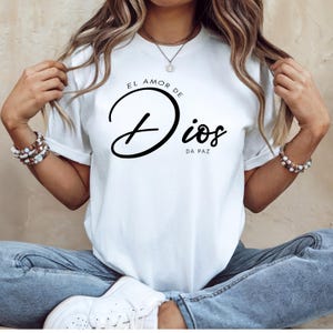 May include: White t-shirt with the text "EL AMOR DE DIOS DA PAZ" in black script. The shirt has a crew neck and short sleeves. The text is centered on the front of the shirt.