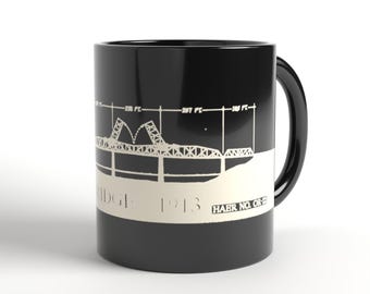 Könnte beinhalten: Ein schwarzer Keramikbecher mit einem weißen Grafikdruck einer architektonischen Brückenskizze. Das Design enthält technische Maße in Fuß, den Text BRIDGE 1913 und HAER NO. OR-22. Der Becher hat einen zylindrischen Körper und einen Henkel.