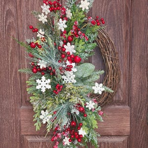May include: A snowy Christmas wreath with red berries, white snowflakes, and green pine branches. The wreath is made of natural twigs and is decorated with festive accents.