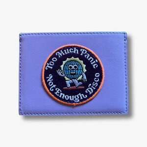 May include: A lavender leather wallet with a circular embroidered patch that reads "Too Much Panic Not Enough Disco" and features a cartoon character with a blue face and a yellow sun.